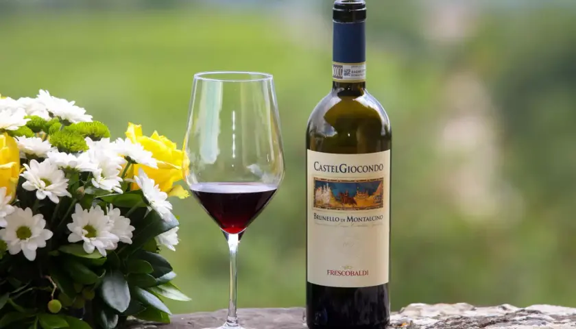 Un bicchiere di vino rosso accanto a una bottiglia di Brunello di Montalcino, con un bouquet di fiori bianchi e gialli.