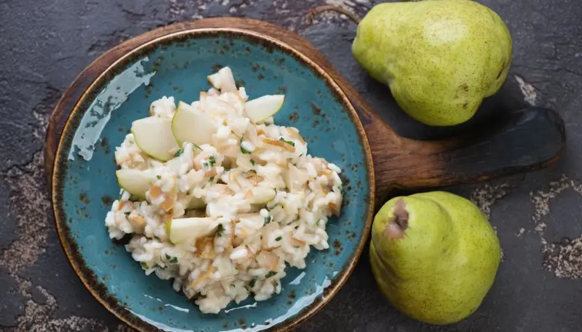 Risotto pere e taleggio cremoso, guarnito con spicchi di pera fresca. Due pere verdi riposano su un tagliere di legno scuro.