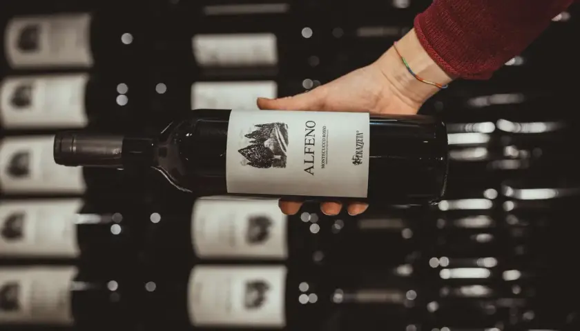 Mano che tiene una bottiglia di vino rosso "Alfero Montecucco Rosso". Il Cabernet Sauvignon, di cui questa regione è originaria, è un vitigno internazionale.