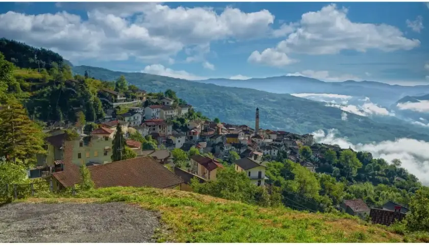 Un borgo di Pregola aggrappato alla collina, con case colorate e un campanile che svetta tra il verde. Nuvole basse accarezzano le montagne sullo sfondo.