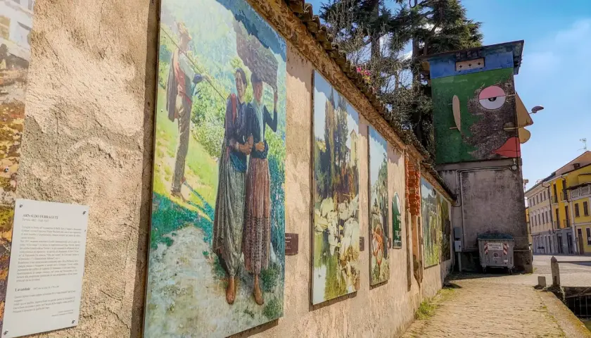 Opere d'arte lungo un muro a Gravellona Lomellina, con dipinti che raffigurano scene di vita rurale. Un'installazione artistica colorata completa il panorama.