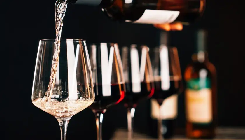 Versando vino bianco in un calice, con altri calici pieni di un intenso rosso rubino sullo sfondo.