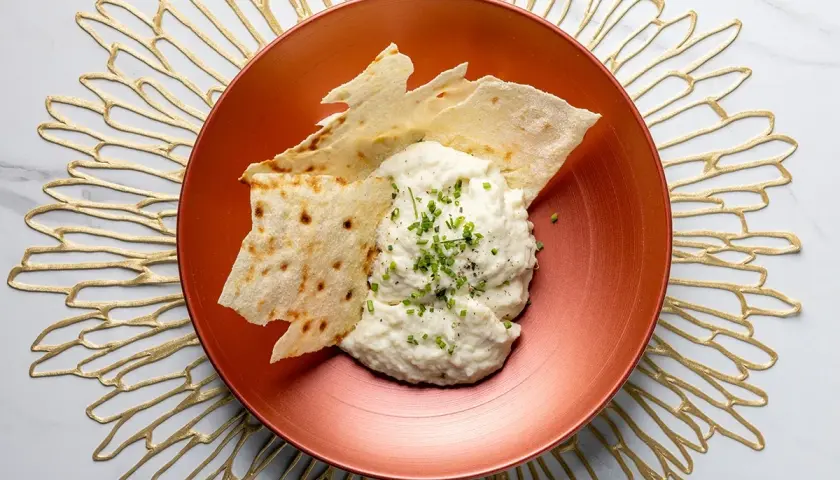 Brandade di baccalà cremosa, guarnita con erba cipollina e pepe, servita con croccanti sfoglie di pane su un piatto ramato.