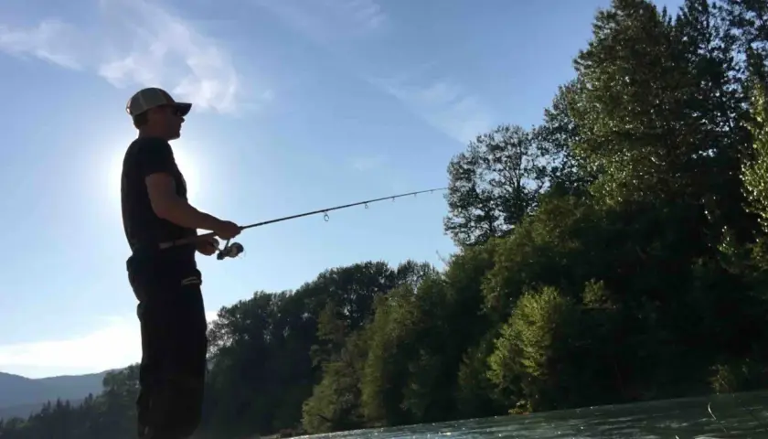 Uomo con cappellino pesca sul fiume Ticino, circondato da alberi e montagne. Un luogo ideale dove pescare sul Ticino.