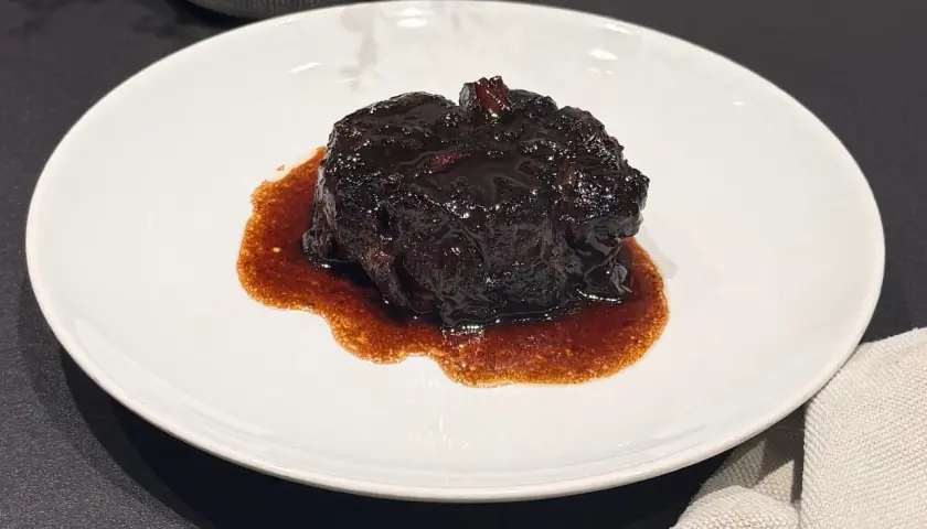 Un succulento brasato al buttafuoco, lucido e scuro, adagiato su un piatto bianco con un sugo denso e aromatico.