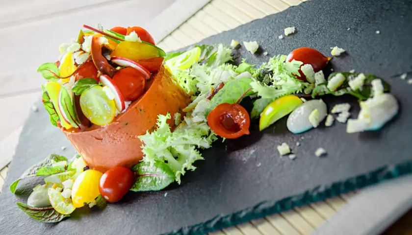 Un'insalata gourmet con terrina arancione, pomodorini colorati, ravanelli, lattuga e una salsa cremosa su una lavagna scura.