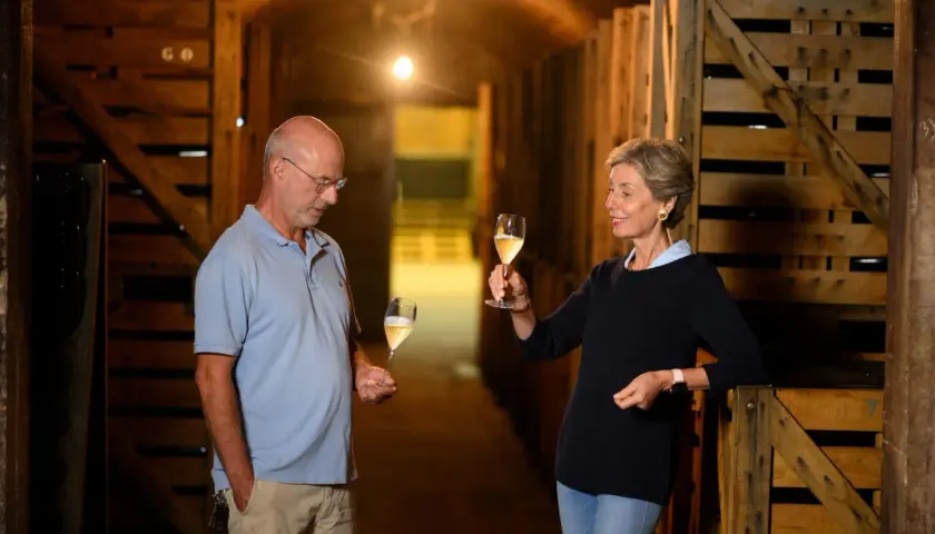 Coppia degusta vino in cantina, tra casse di legno. Il vino, affinato sur lie, brilla nei calici.