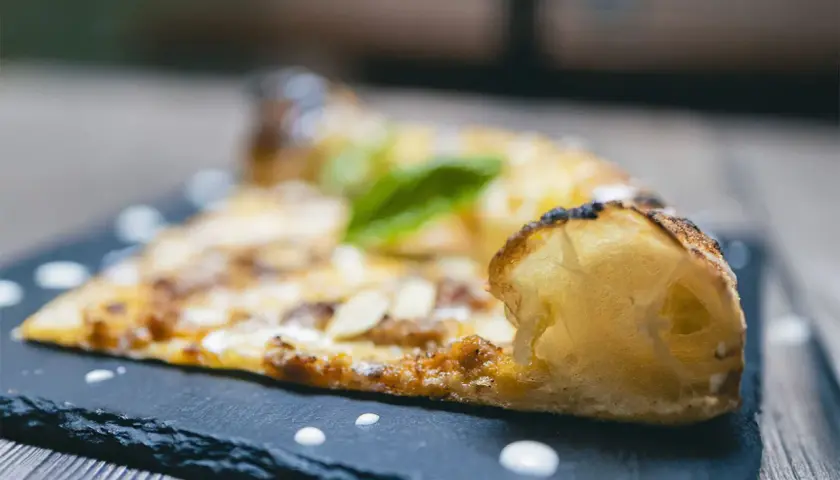 Una fetta di pizza al taglio con mandorle e basilico, servita su una lavagna scura con gocce di salsa.