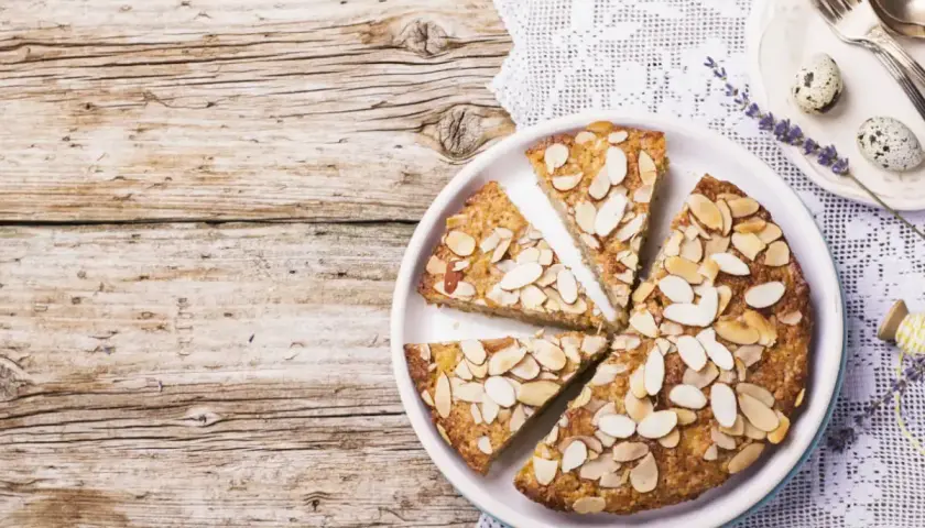 Fetta di torta di mandorle su un piatto bianco, guarnita con mandorle a lamelle. Sullo sfondo, un tovagliolo di pizzo e due uova di quaglia.