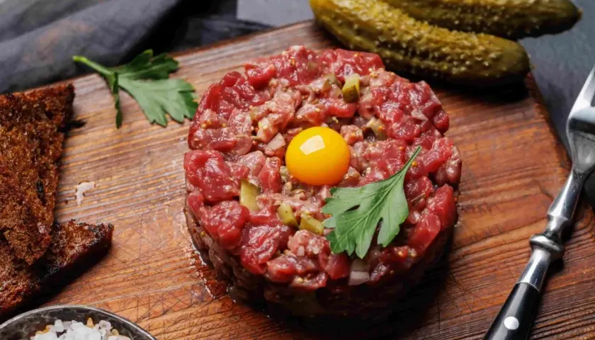 Tartare di carne cruda, con tuorlo d'uovo, cetriolini, pane tostato, sale grosso e prezzemolo su tagliere di legno.