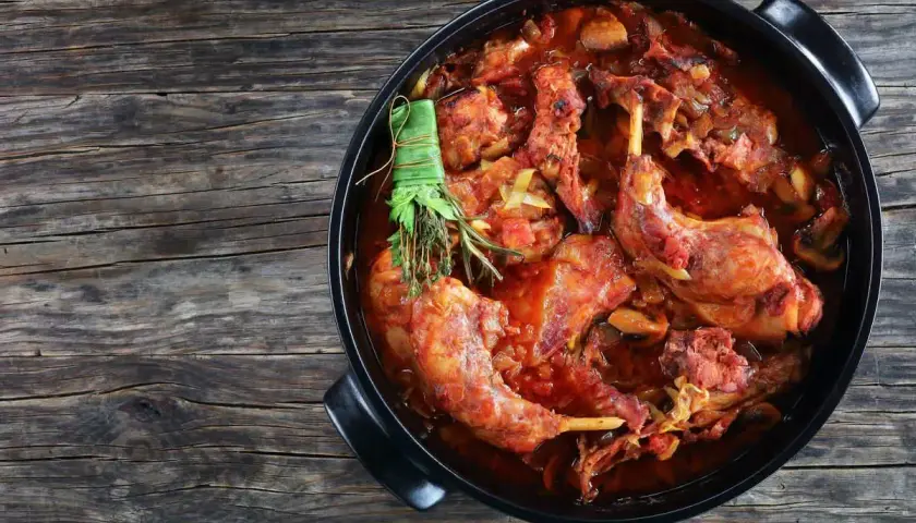 Un saporito coniglio al vino rosso, cotto lentamente con erbe aromatiche, in una casseruola nera su un tavolo di legno rustico.