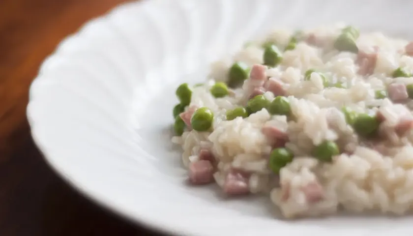 Un cremoso riso con piselli e cubetti di prosciutto, servito su un piatto bianco con un bordo ondulato.