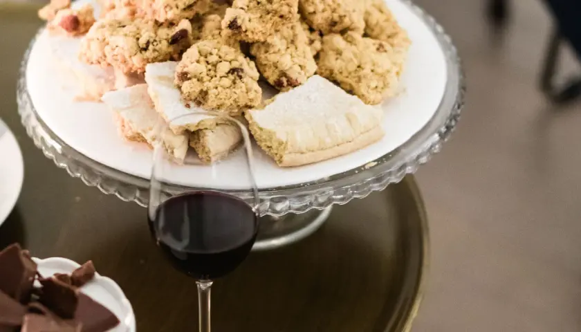 A glass of red wine sits next to a cake stand filled with cookies and pastries.