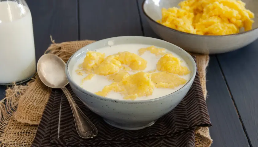 Una ciotola di polenta e latte, un piatto confortante. Accanto, una bottiglia di latte e un cucchiaio d'argento.