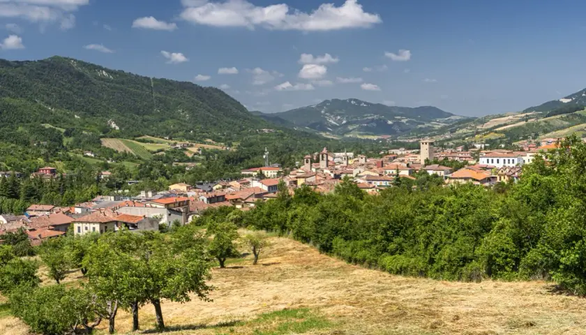 Panoramica del borgo del monte antico Varzi, con case dai tetti rossi incastonate tra colline verdi e alberi rigogliosi sotto un cielo azzurro.