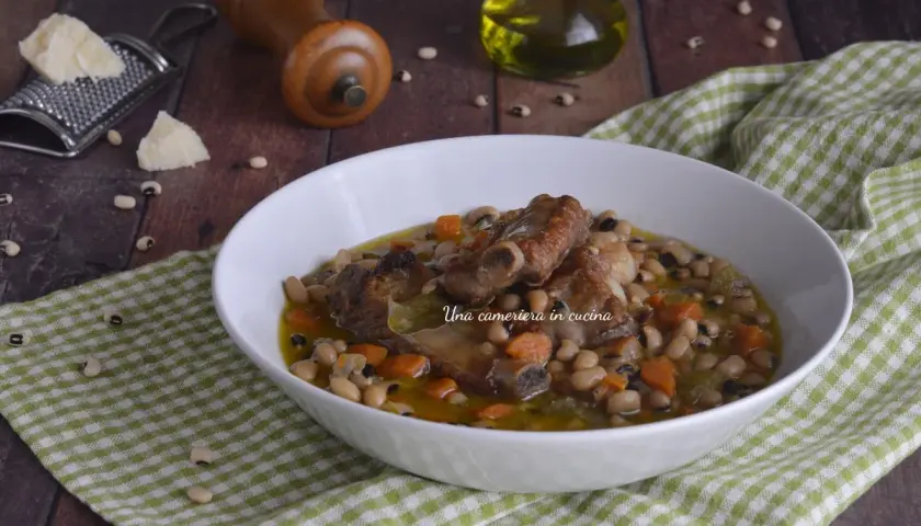 Una ciotola di zuppa dei morti con fagioli, costine di maiale e carote, servita su una tovaglia a quadri verdi.