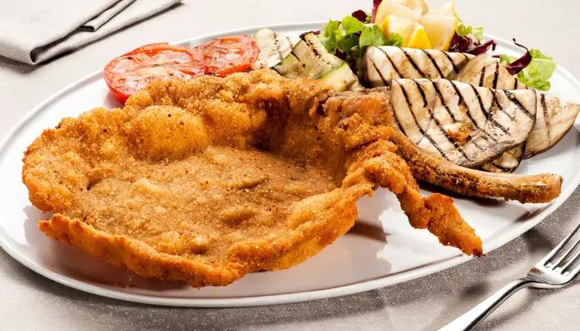Cotoletta alla milanese dorata e croccante, servita con pomodori grigliati, melanzane e un'insalata fresca.