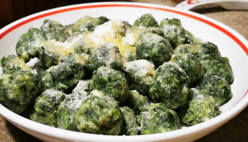 Malfatti ricetta originale: gnocchetti verdi fatti in casa, conditi con burro fuso e parmigiano grattugiato, serviti in una ciotola bianca con bordo rosso.