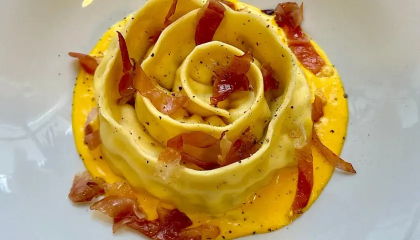 Pappardella ripiena a forma di rosa, adagiata su una crema gialla e guarnita con croccante prosciutto.