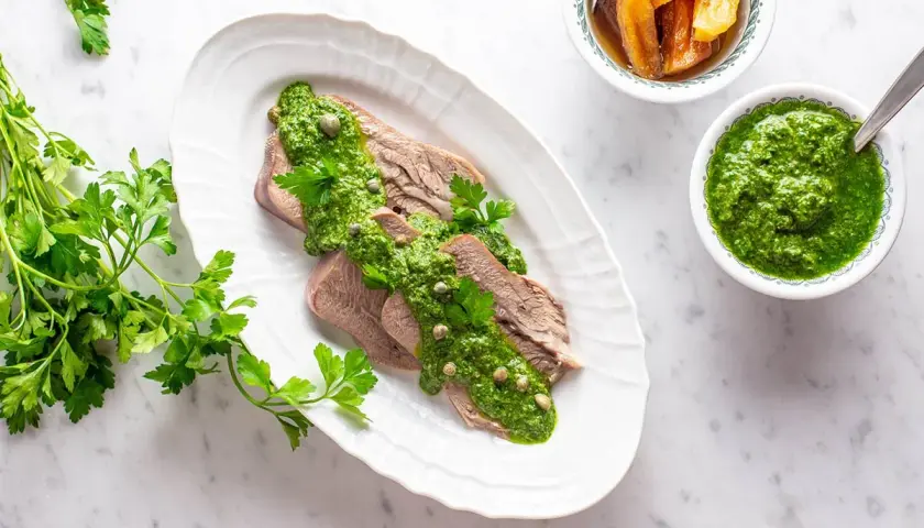 Lingua bollita affettata, generosamente condita con salsa verde e capperi. Accanto, una ciotola di salsa verde e altra guarnizione.