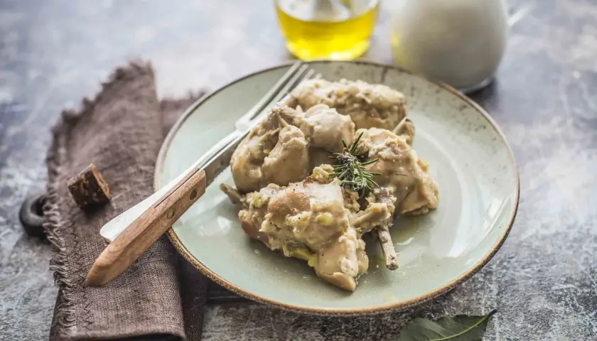 Piatto di coniglio al latte, tenero e saporito, guarnito con rosmarino. Accanto, olio e latte in caraffa.