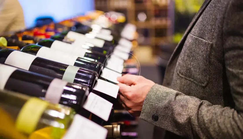 Un uomo in giacca sceglie una bottiglia di vino, forse un pregiato vino muffato, tra una fila di etichette bianche.