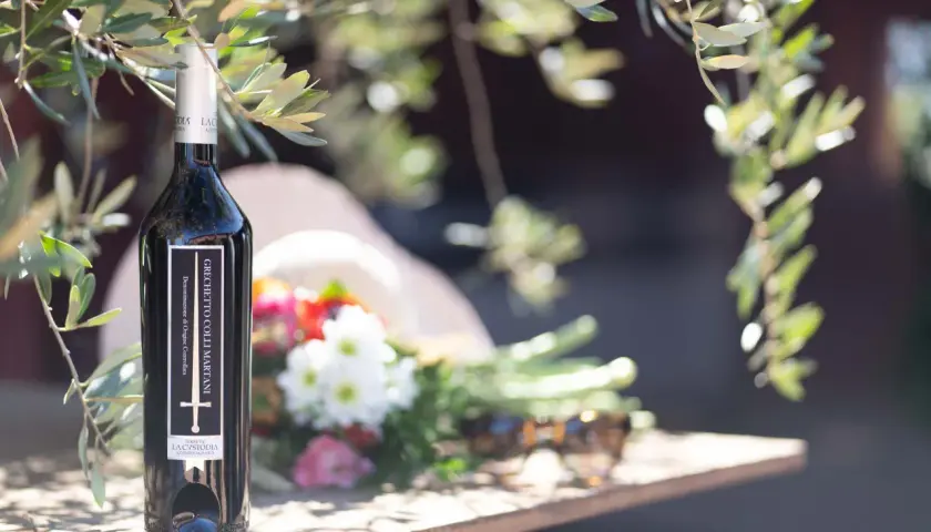 Bottiglia di vino Grechetto Colli Martani, con fiori e occhiali da sole su un tavolo di legno, sotto i rami di un ulivo.
