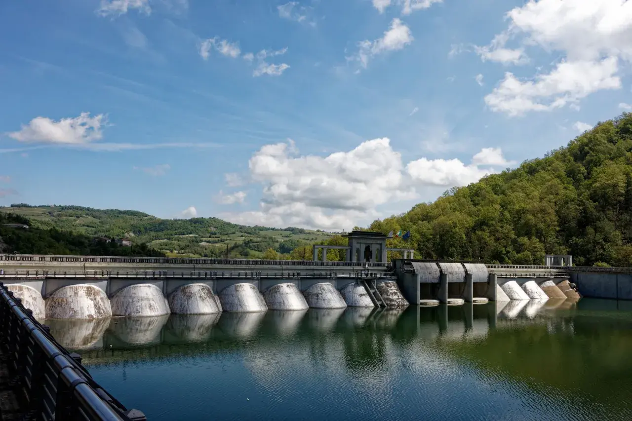 Diga imponente con colline verdi sullo sfondo, ideale per il trekking a Pavia. L'acqua riflette il cielo azzurro e le nuvole bianche.