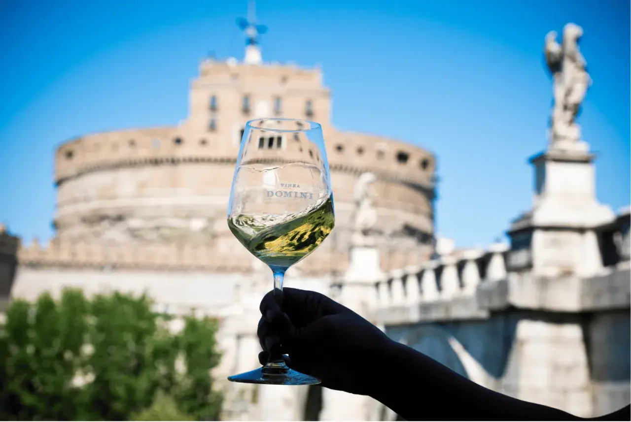 Un calice di vino bianco ondeggia con il logo "Vinea Domini" sullo sfondo di Castel Sant'Angelo a Roma.