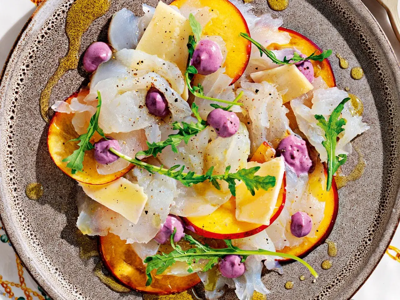 Delicato carpaccio di pesce, fettine di pesca, formaggio, rucola e sfere viola, un'interpretazione moderna dei frutti di mare crudi.