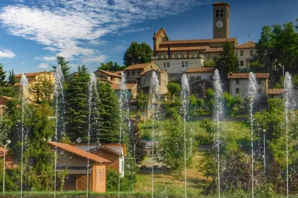 Fontane zampillanti davanti a un borgo collinare con chiesa e campanile. Un'immagine che invita a scoprire cosa vedere a Fortunago.