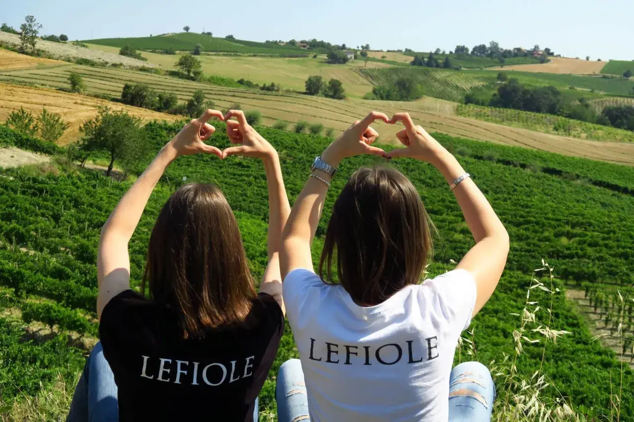 Due amiche formano cuori con le mani, con sullo sfondo le colline di Redavalle e i vigneti che daranno vita al vino.