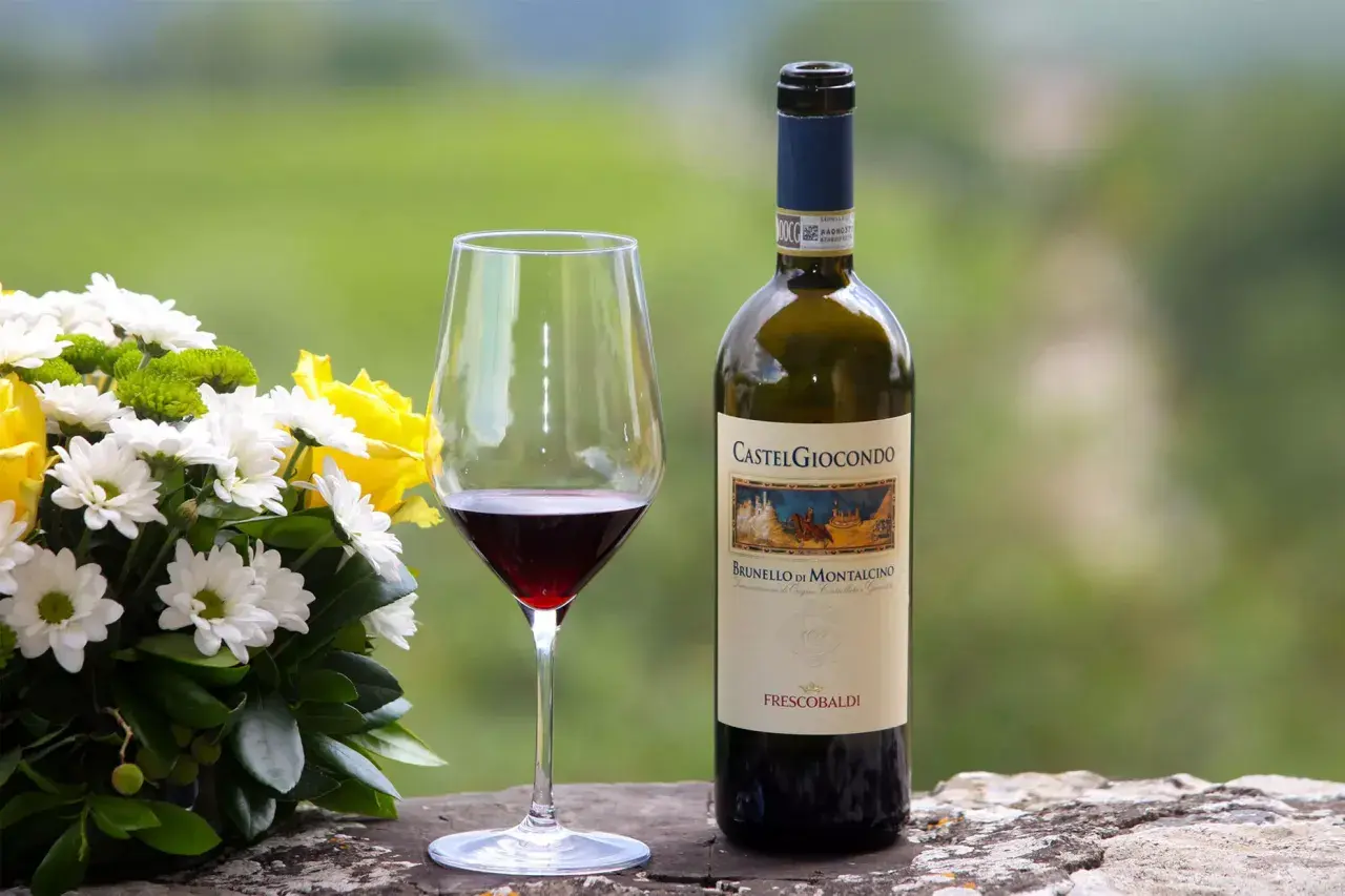 Un bicchiere di vino rosso accanto a una bottiglia di Brunello di Montalcino, con un bouquet di fiori bianchi e gialli.