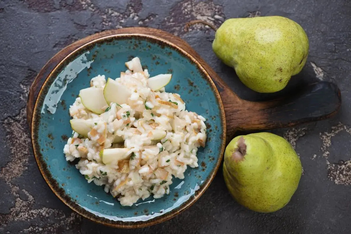Risotto pere e taleggio cremoso, guarnito con spicchi di pera fresca. Due pere verdi riposano su un tagliere di legno scuro.