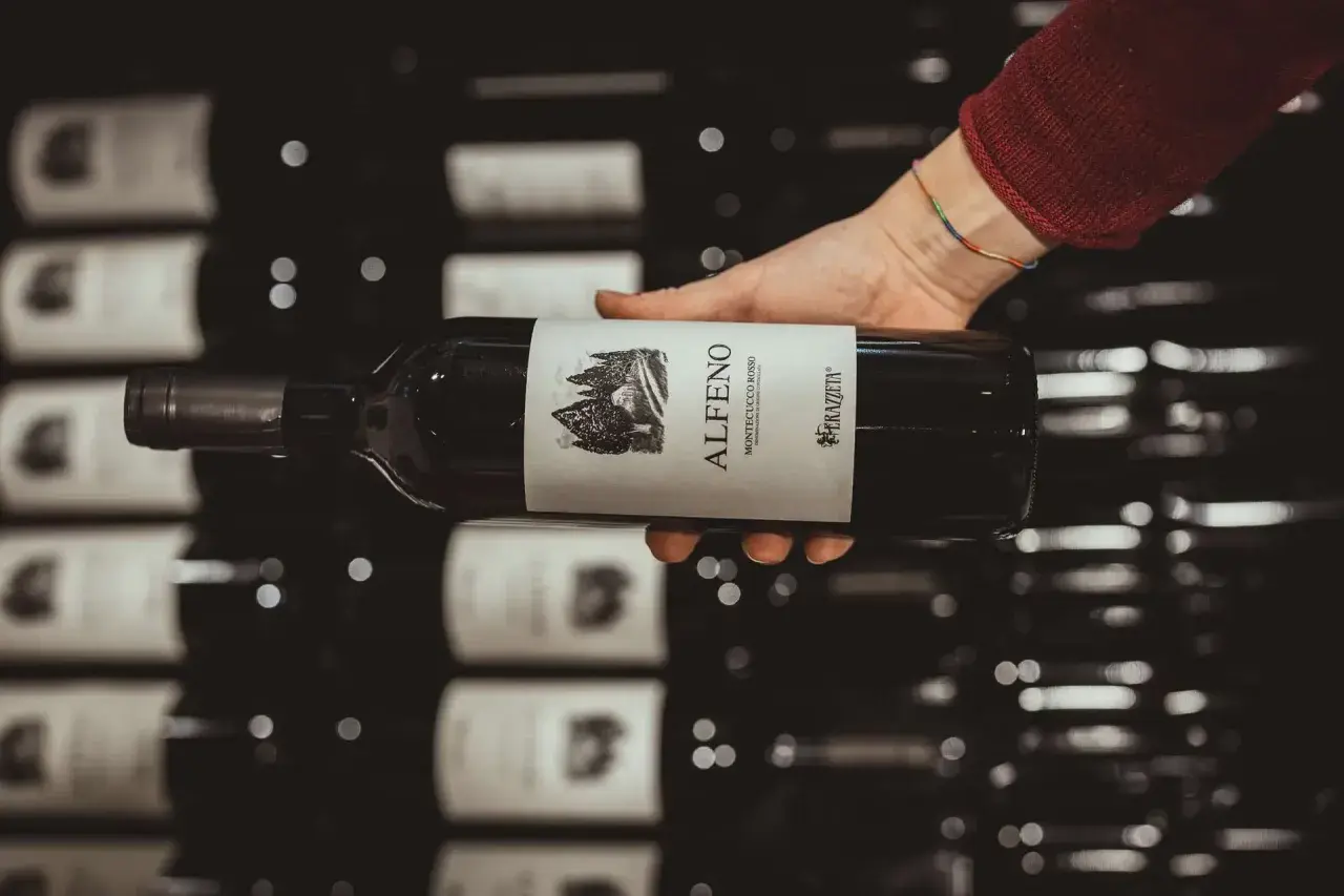 Mano che tiene una bottiglia di vino rosso "Alfero Montecucco Rosso". Il Cabernet Sauvignon, di cui questa regione è originaria, è un vitigno internazionale.