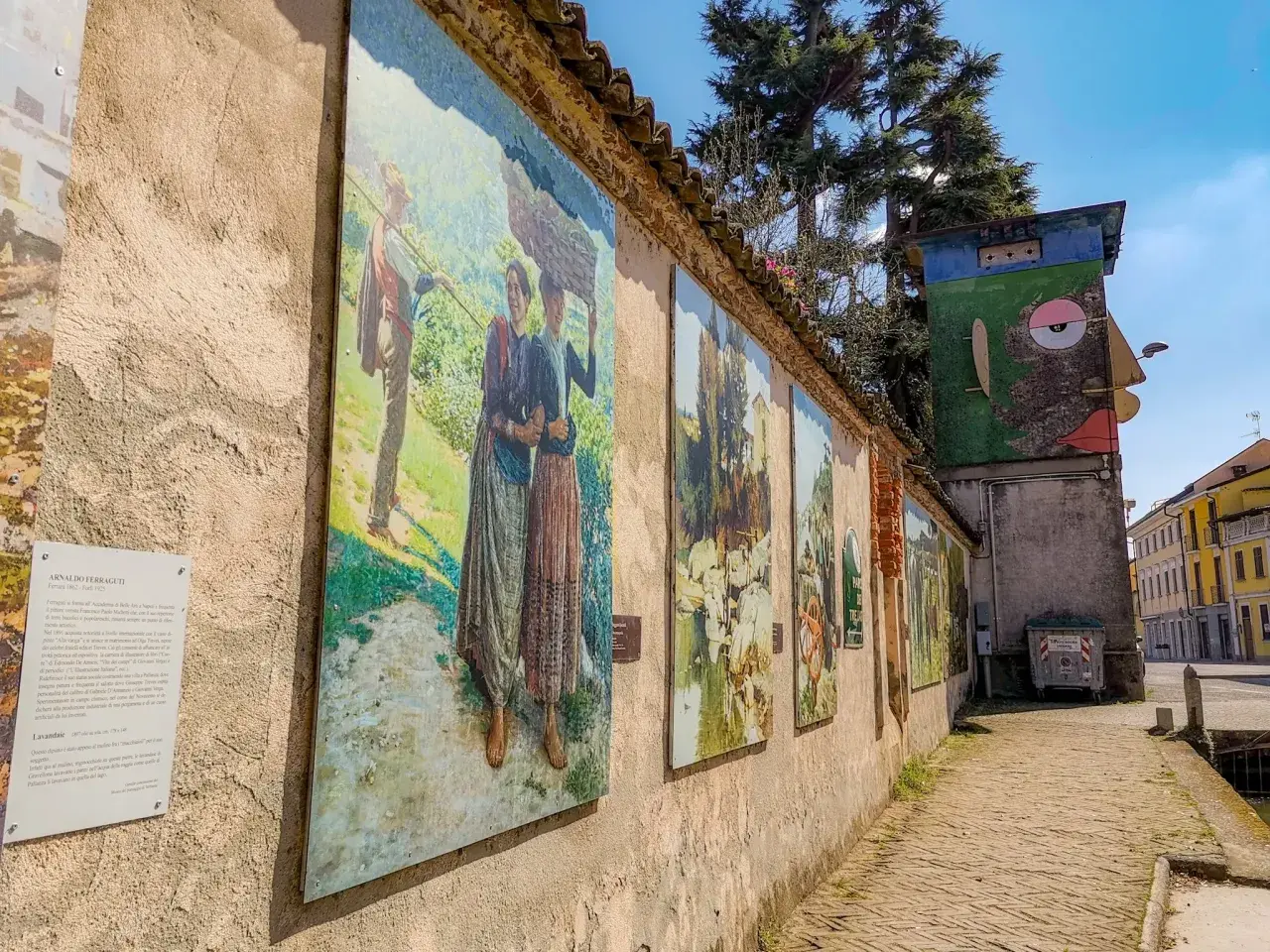 Opere d'arte lungo un muro a Gravellona Lomellina, con dipinti che raffigurano scene di vita rurale. Un'installazione artistica colorata completa il panorama.