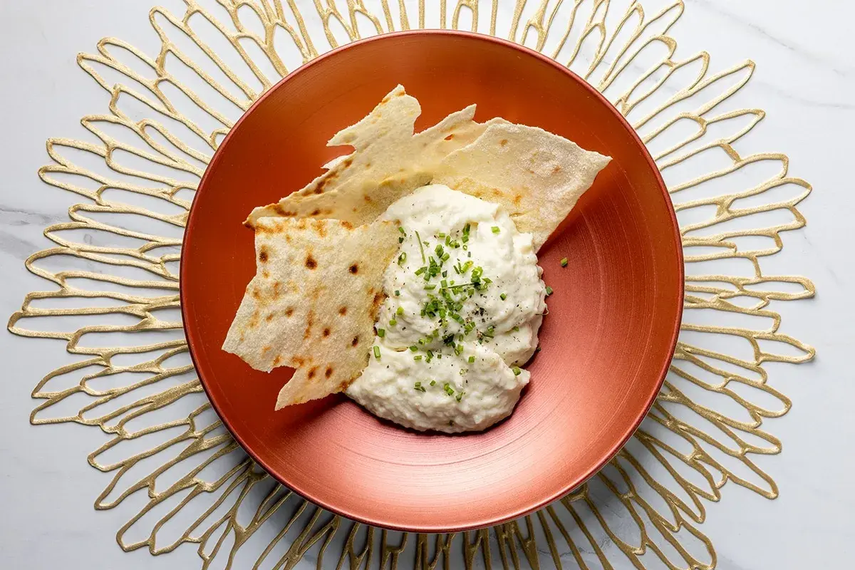 Brandade di baccalà cremosa, guarnita con erba cipollina e pepe, servita con croccanti sfoglie di pane su un piatto ramato.