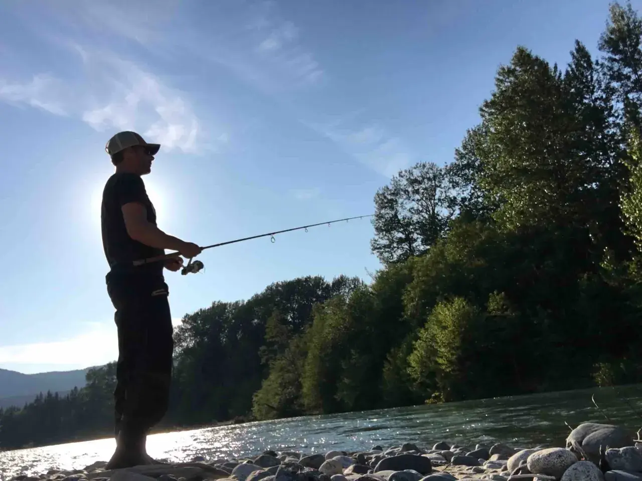 Uomo con cappellino pesca sul fiume Ticino, circondato da alberi e montagne. Un luogo ideale dove pescare sul Ticino.