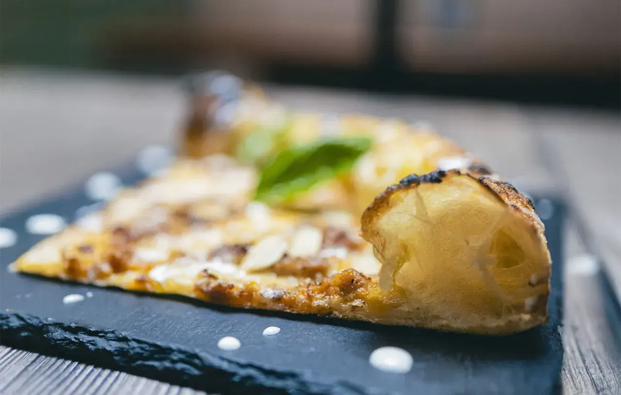 Una fetta di pizza al taglio con mandorle e basilico, servita su una lavagna scura con gocce di salsa.