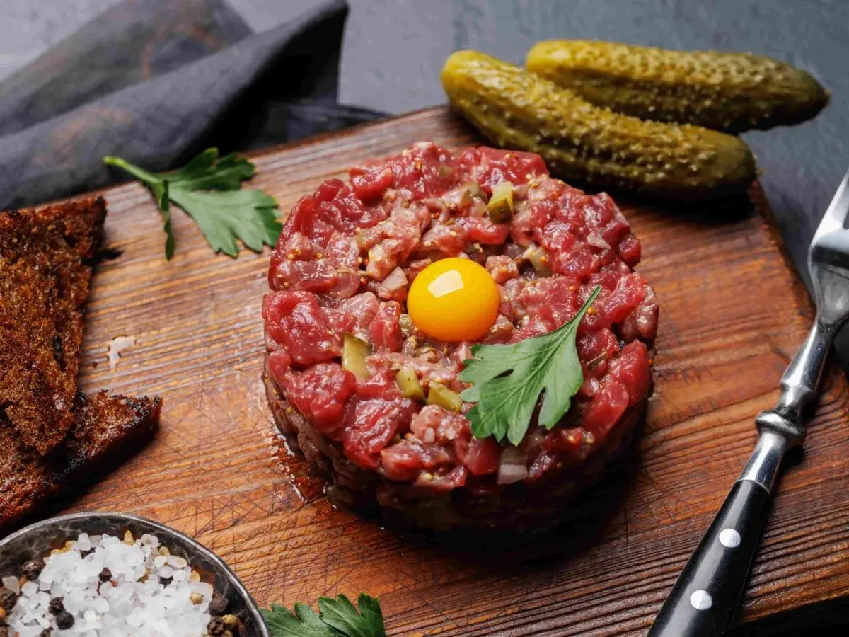 Tartare di carne cruda, con tuorlo d'uovo, cetriolini, pane tostato, sale grosso e prezzemolo su tagliere di legno.