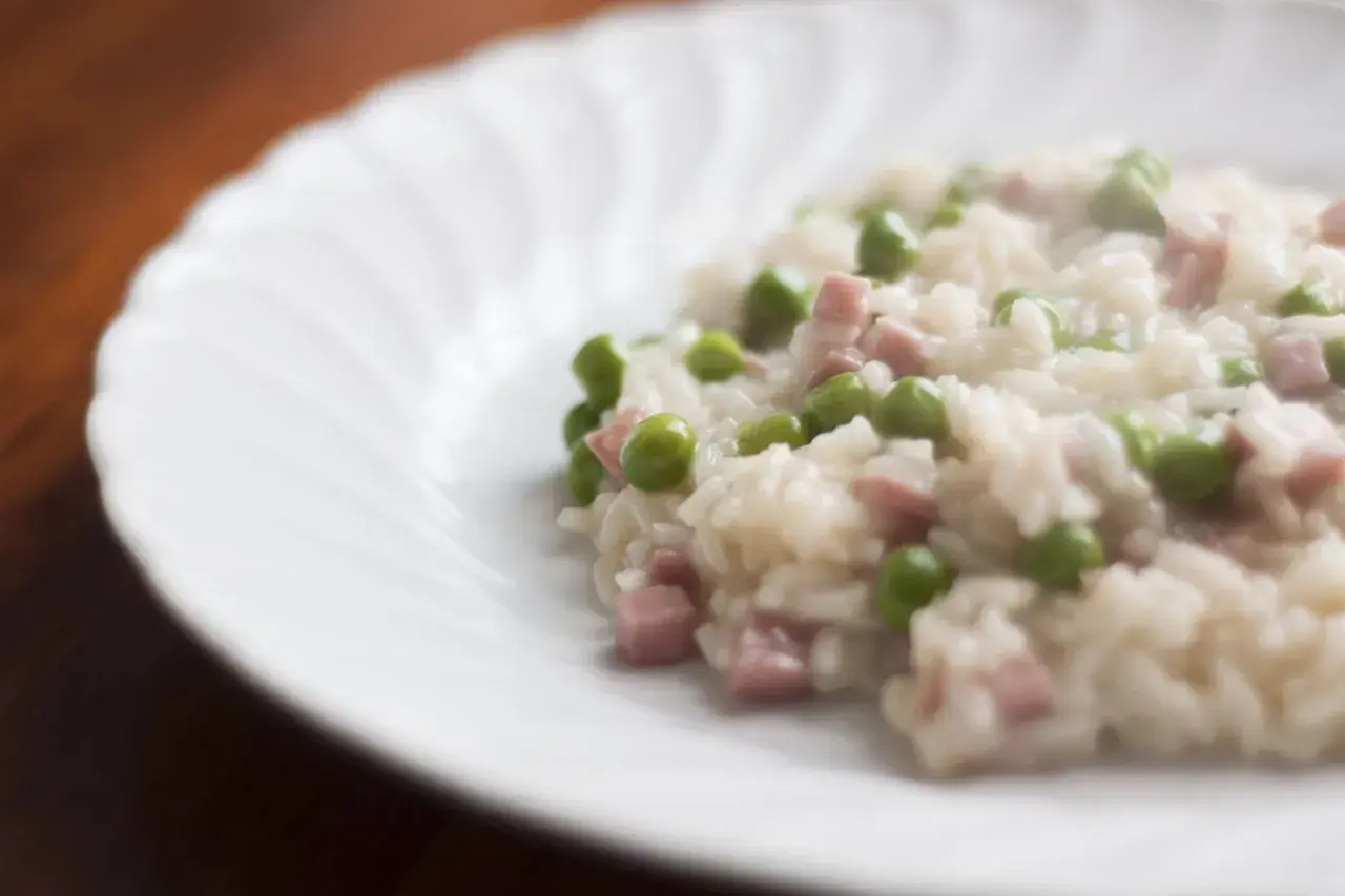 Un cremoso riso con piselli e cubetti di prosciutto, servito su un piatto bianco con un bordo ondulato.