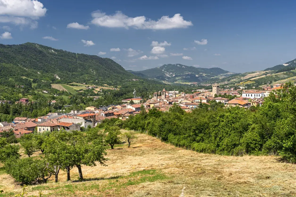 Panoramica del borgo del monte antico Varzi, con case dai tetti rossi incastonate tra colline verdi e alberi rigogliosi sotto un cielo azzurro.
