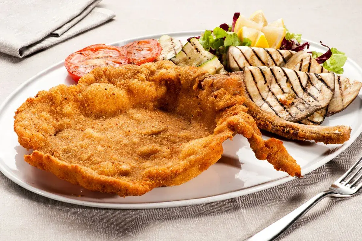 Cotoletta alla milanese dorata e croccante, servita con pomodori grigliati, melanzane e un'insalata fresca.