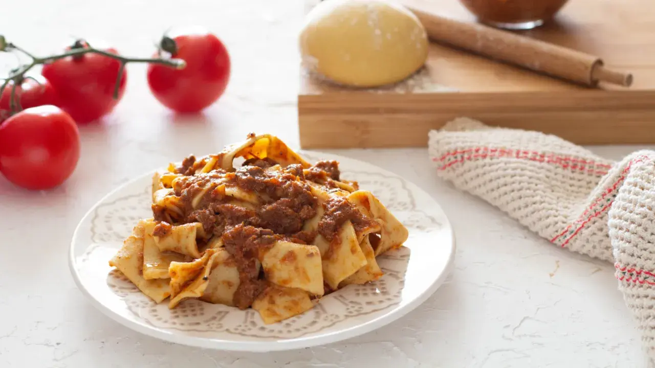 Piatto di pappardelle alla lepre, condite con un ricco ragù. Sullo sfondo, pomodori freschi, impasto e mattarello.