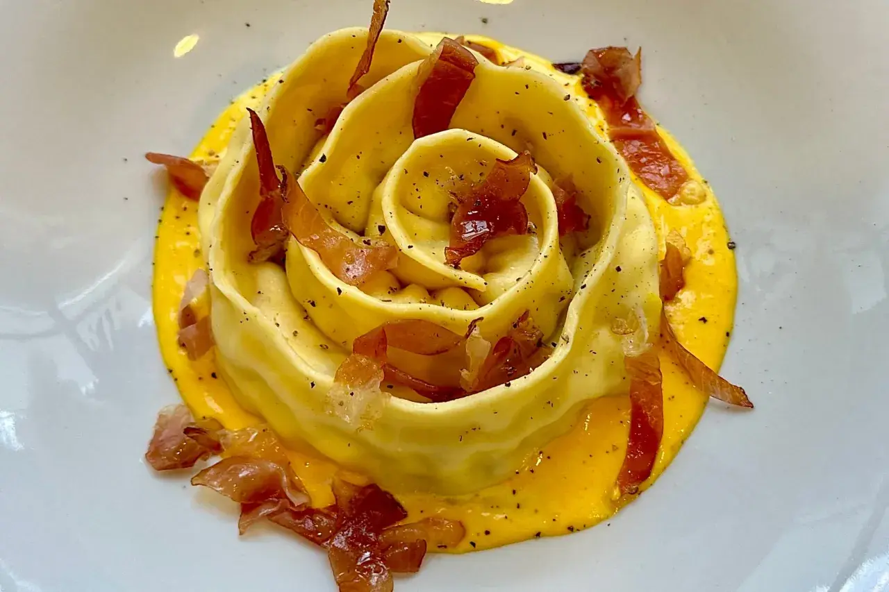 Pappardella ripiena a forma di rosa, adagiata su una crema gialla e guarnita con croccante prosciutto.