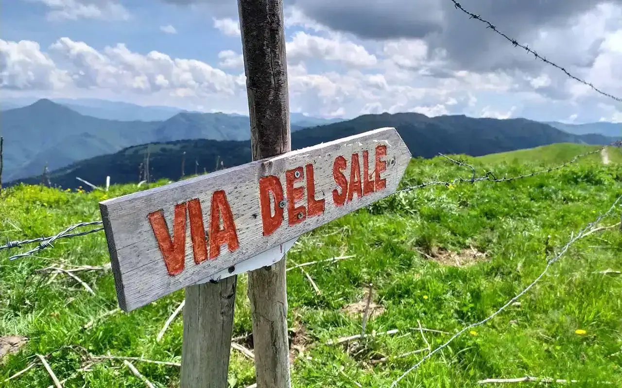 Cartello in legno "Via del Sale Varzi" su un sentiero montano verdeggiante, con cielo nuvoloso e montagne sullo sfondo.