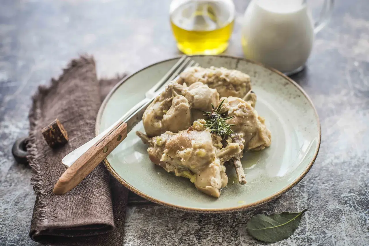Piatto di coniglio al latte, tenero e saporito, guarnito con rosmarino. Accanto, olio e latte in caraffa.