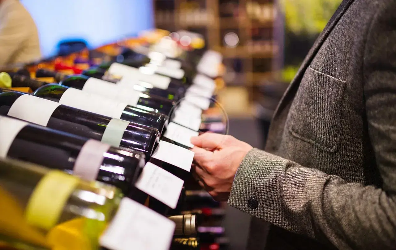 Un uomo in giacca sceglie una bottiglia di vino, forse un pregiato vino muffato, tra una fila di etichette bianche.