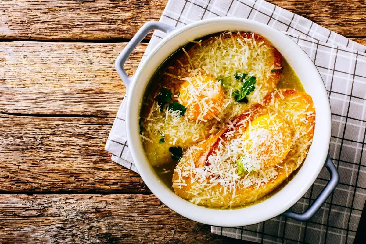 Zuppa alla pavese fumante, con pane tostato, uova e abbondante parmigiano grattugiato, servita in una ciotola bianca su un tavolo di legno rustico.