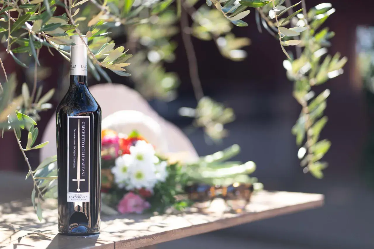 Bottiglia di vino Grechetto Colli Martani, con fiori e occhiali da sole su un tavolo di legno, sotto i rami di un ulivo.